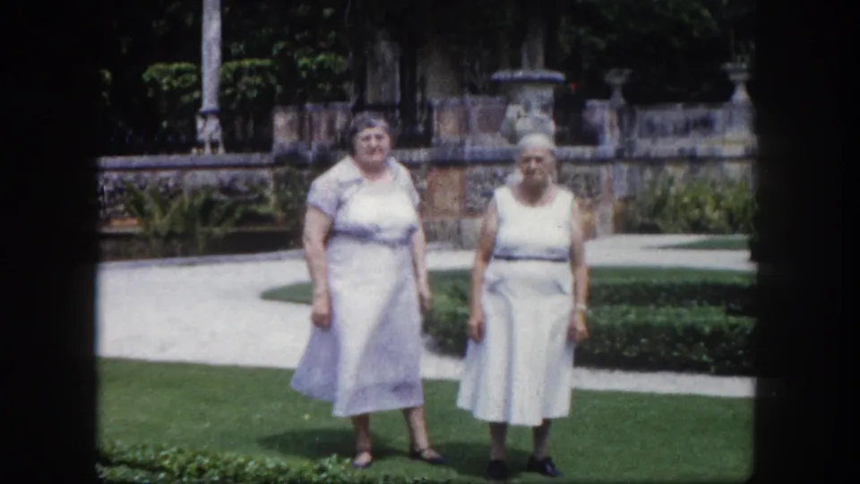Two older women in early-1960s pale dresses standing on a lawn in a formal Tampa garden, ornate stone balustrade with decorative urns and tropical trees visible behind them.