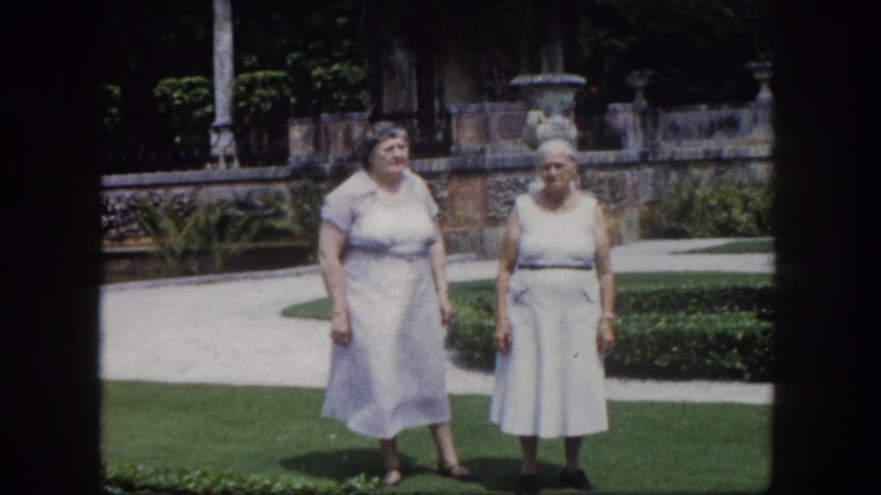 Two older women in pale 1960s dresses standing on a formal Florida garden lawn; stone urns on a balustrade and lush tropical foliage visible in strong sunlight.