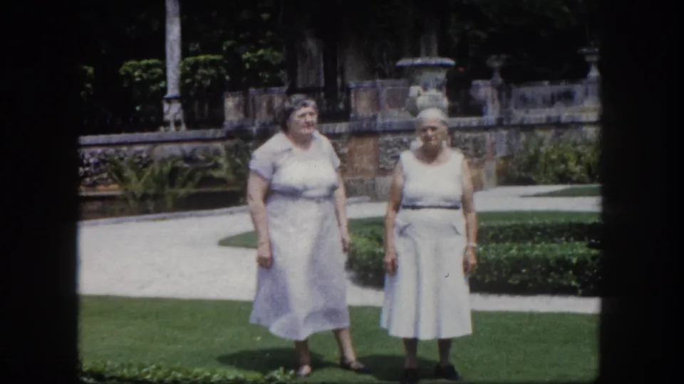 Two older women in pale 1960s dresses standing on a formal Florida garden lawn; stone urns on a balustrade and lush tropical foliage visible in strong sunlight.