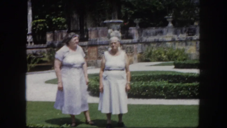Two older women in pale 1960s dresses, one beginning to turn, in a sunlit Florida formal garden; stone balustrade with ornamental urns prominent in background.
