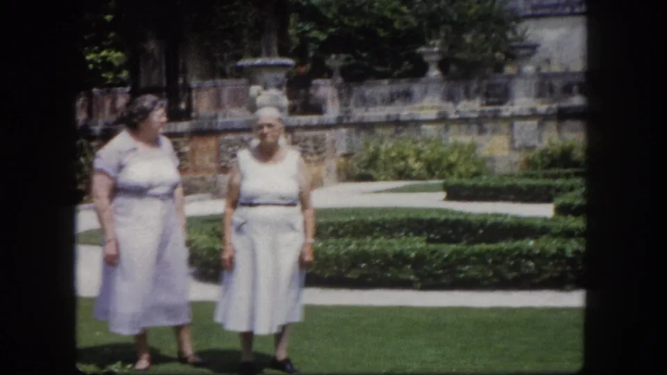 Two older women in near-profile view on a formal Florida garden lawn circa 1960, ornate stone urns and hedgerow visible in bright sunlight.