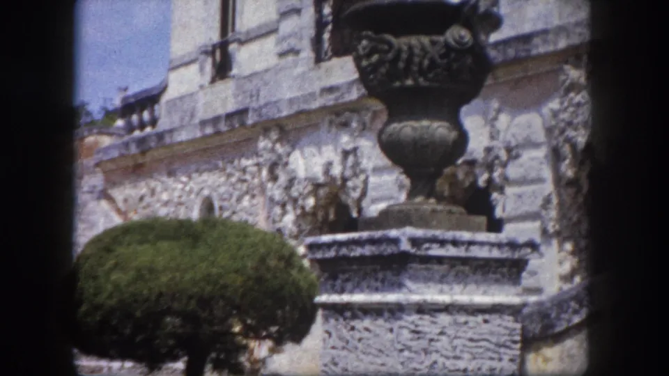 Large ornate cast-metal urn with scroll and shell relief on a stone pedestal, carved stone building wall with relief panels behind, Tampa, Florida, circa 1960.