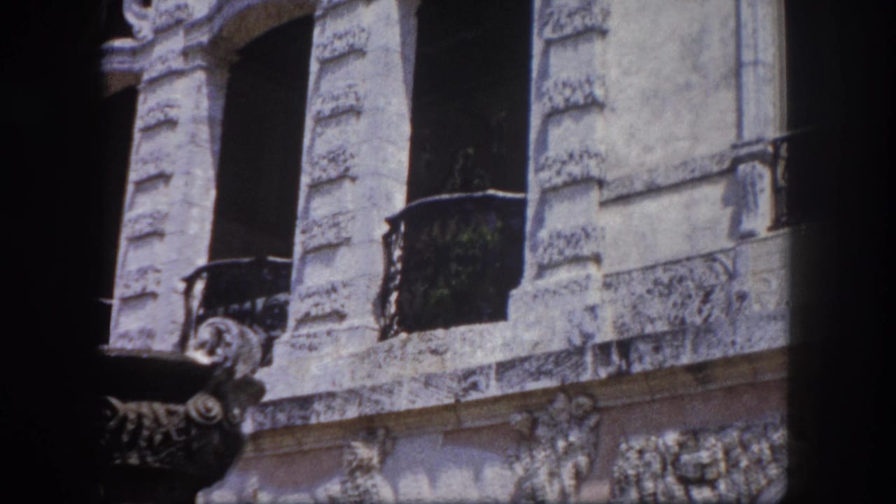 Ornate multi-story stone building facade with arched windows and wrought iron balconies; a large classical decorative urn appears at right foreground; 8mm, Tampa, Florida, 1960.
