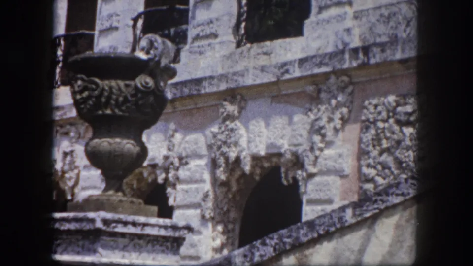 Large ornate classical urn on a stone pedestal dominates the foreground; multi-story carved stone building facade with arched windows and wrought iron balconies behind; 8mm, Tampa, Florida, 1960.