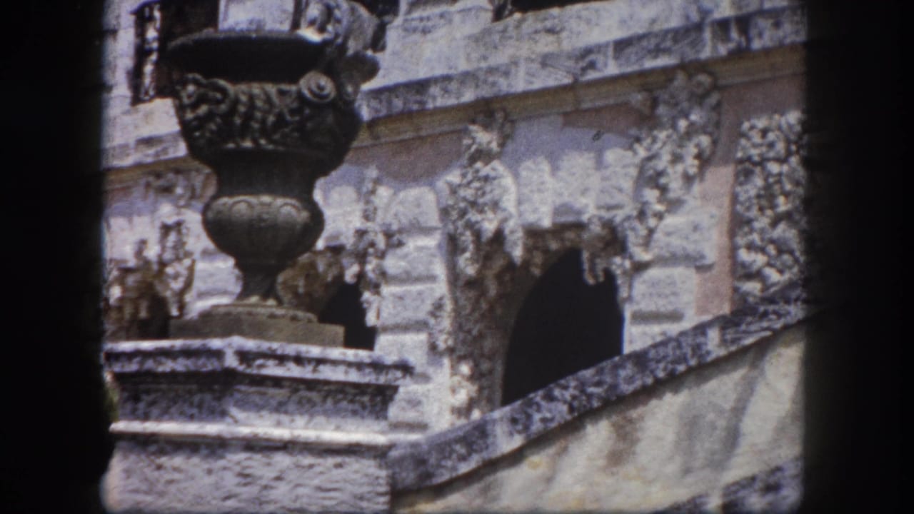 Two ornate stone urns on carved pedestals flank a relief-carved stone facade with balustraded steps, framed by 8mm film gate borders.