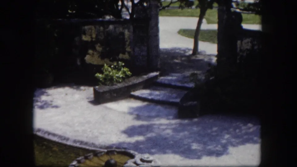 Sunlit estate courtyard with stone steps, low stone water feature, garden path, and trimmed trees, Tampa Florida area, circa 1960 8mm footage.