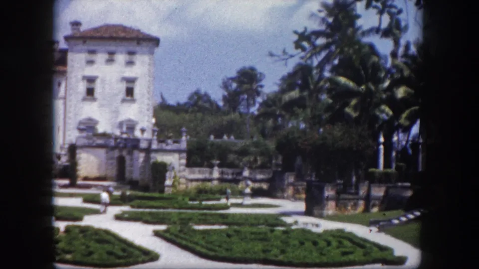 Florida estate exterior with formal garden hedges, stone balustrades, tropical palms, and small distant visitors on garden paths, circa 1960, 8mm color film.