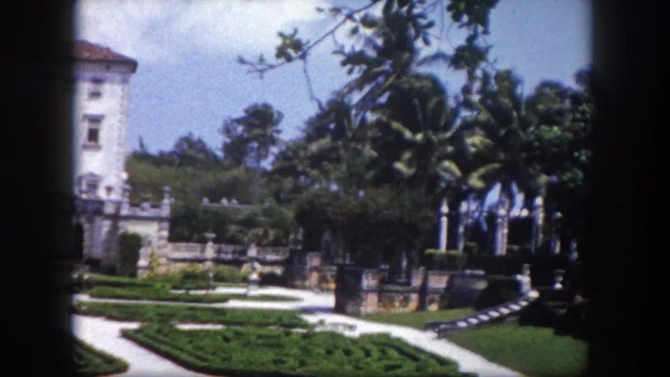 Florida estate building with tropical palms and large foreground tree framing formal garden hedge patterns; similar composition with slight camera shift, circa 1960, 8mm color.