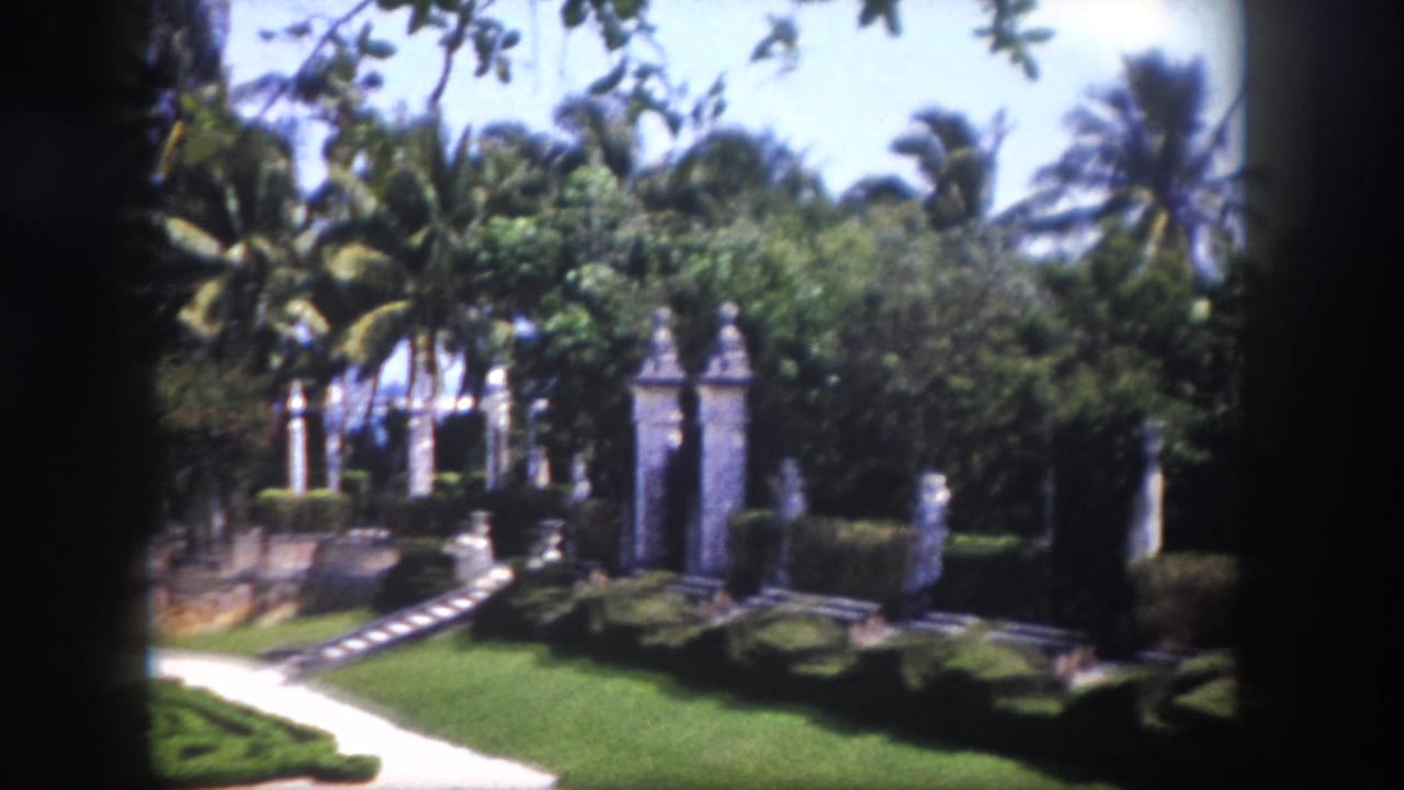 Row of stone pillars beside a formal garden path, manicured lawn and hedges, tropical trees beyond, 8mm film.