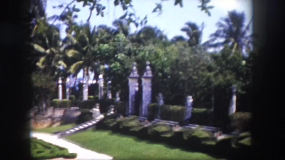 Row of classical stone pillars flanking a formal garden path with clipped hedges and palm trees beyond, Tampa Florida area circa 1960, 8mm film.