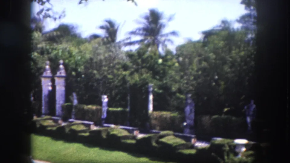 Slow pan across formal garden with classical stone columns, clipped hedges, and tall tropical palms, Florida circa 1960, 8mm film.