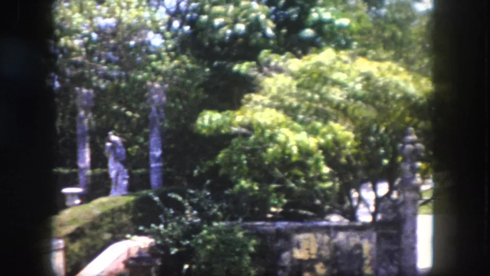 1960 8mm home movie frame of a formal ornamental garden in Tampa, Florida, showing a rectangular reflecting pond and stone gate pillars in bright sunshine.