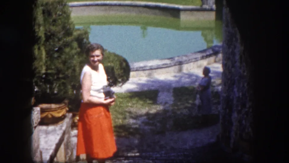 1960 8mm home movie showing a smiling woman in white blouse and orange midi skirt posed beside a formal garden pond in Tampa, Florida.