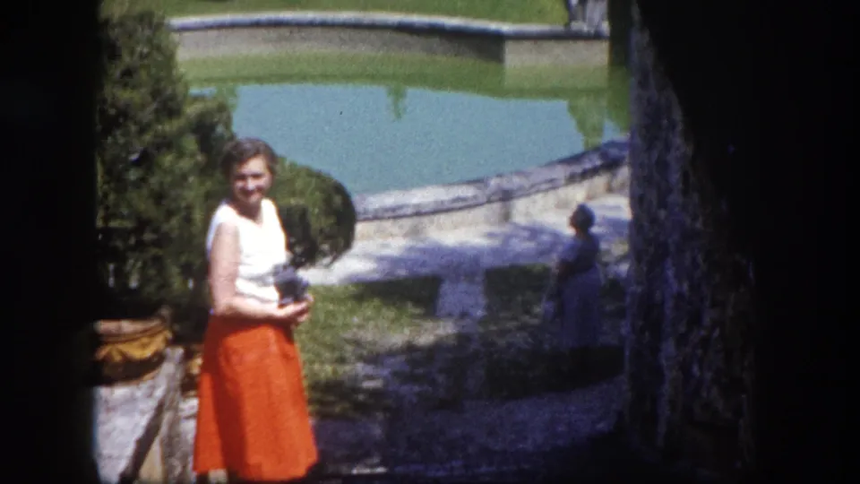 1960 8mm home movie frame of a woman in white blouse and orange skirt smiling beside a formal garden pond, stone balustrade and subtropical trees visible behind her in Tampa, Florida.