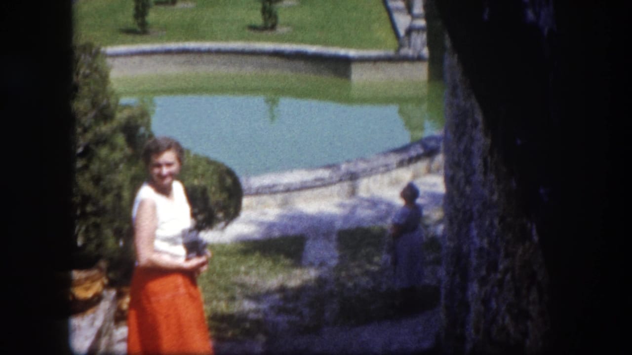 Woman in orange skirt and white top stands smiling before a large reflecting pool with manicured grounds and topiary behind her.