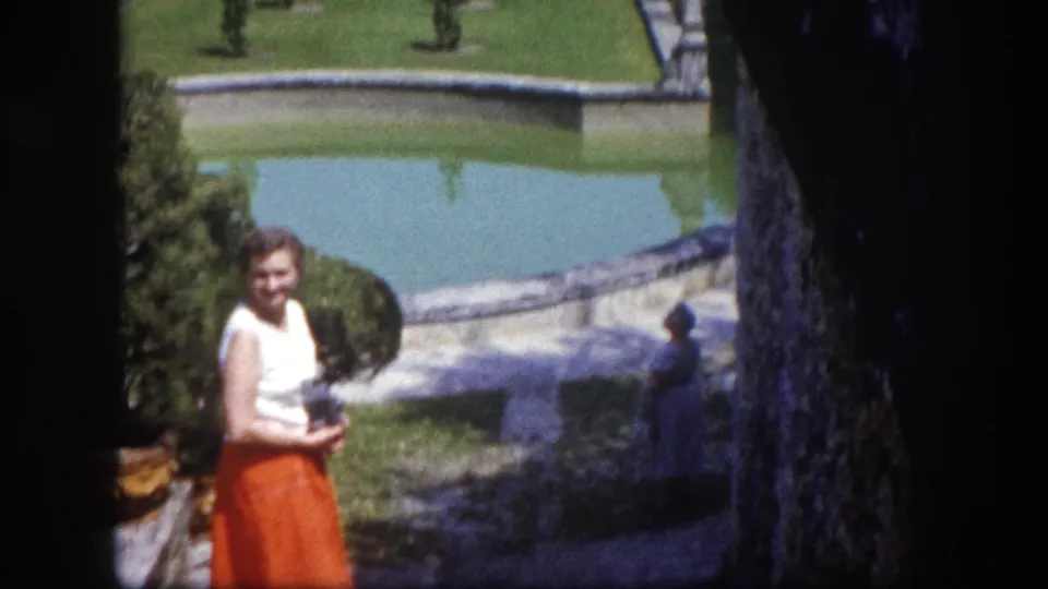 Early-1960s woman in orange midi skirt poses at a formal estate garden with a large reflecting pool and topiary, Tampa, Florida, circa 1960.
