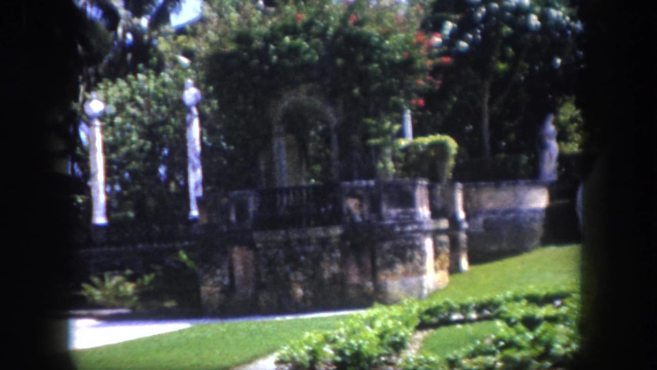 Garden terrace with ornamental white columns, stone arched wall, and tropical trees; large ornate iron finial partially visible at center.