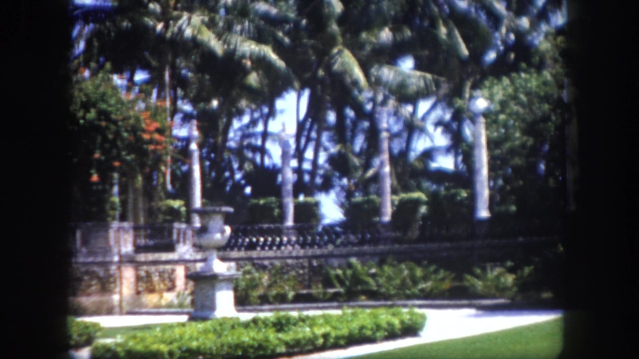 Formal garden with stone balustrade, tall palm trees, and ornate columned gateway; a distant figure is barely visible near the gate.