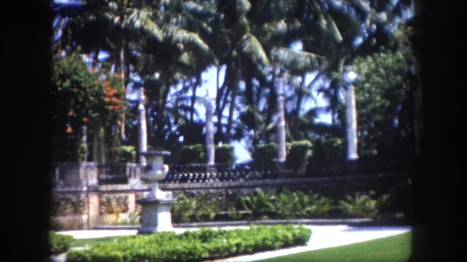 8mm home movie frame circa 1960, Tampa, Florida: formal Mediterranean-style garden with stone balustrade, palm trees, ornate gateway, and a distant figure.