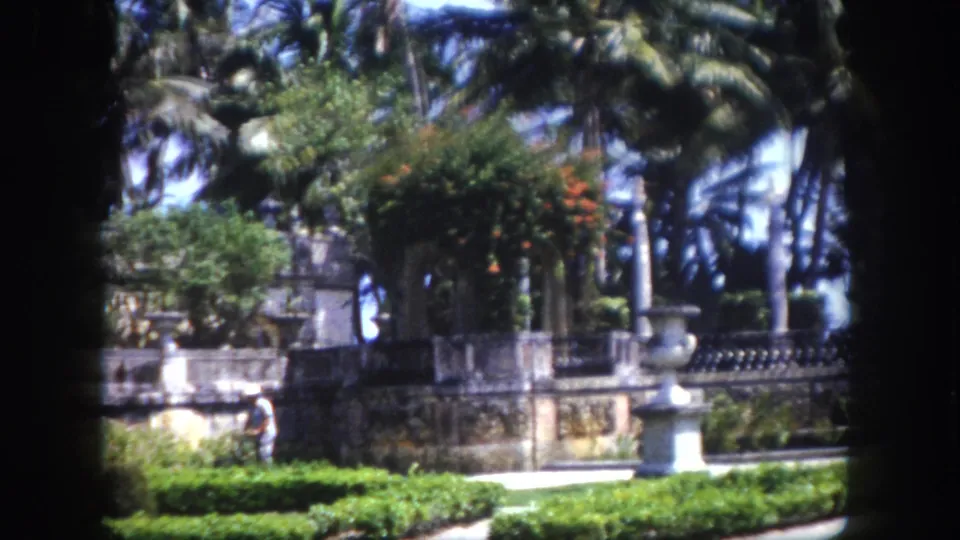 8mm home movie circa 1960, Tampa, Florida: manicured hedges and decorative stone balustrade in foreground, palm trees behind, figure in light clothing walking.