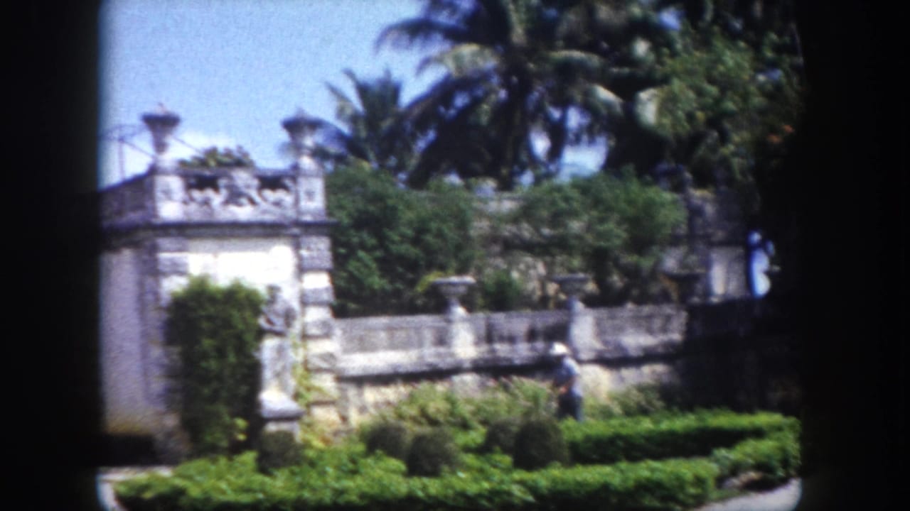 Ornate stone garden wall with classical statues and manicured hedges; a person visible among the plantings.