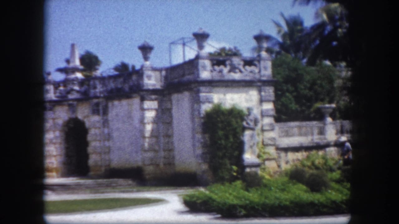 8mm frame circa 1960, Florida: ornate classical stone gate with arched entrance, decorative statues atop balustrade wall, curved driveway and green shrubbery visible.