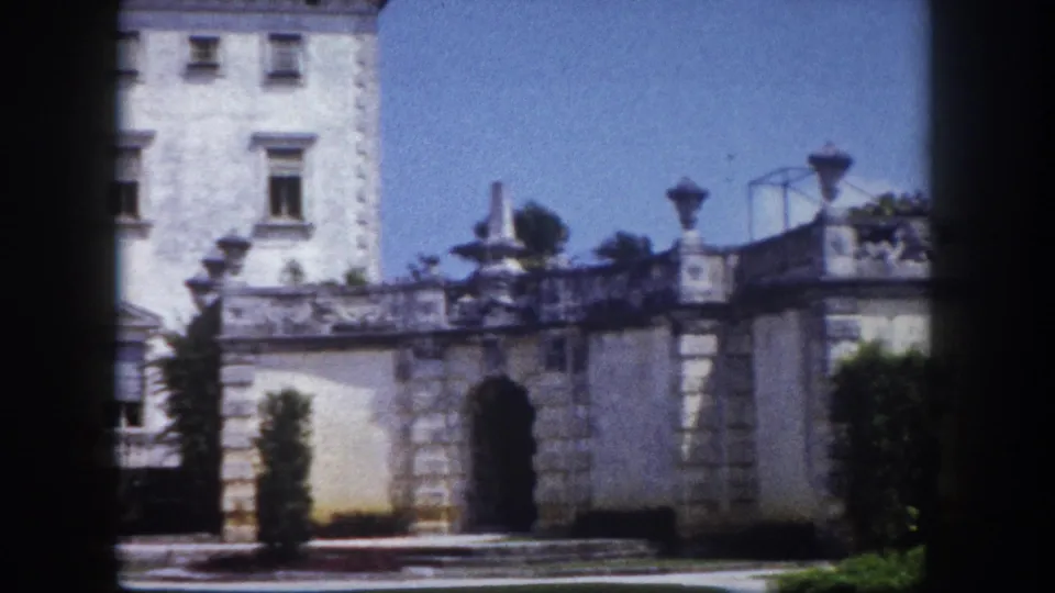 8mm frame circa 1960, Florida: large white multi-story Mediterranean Revival mansion with rows of windows, ornate stone balustrade wall and decorative urns in foreground.