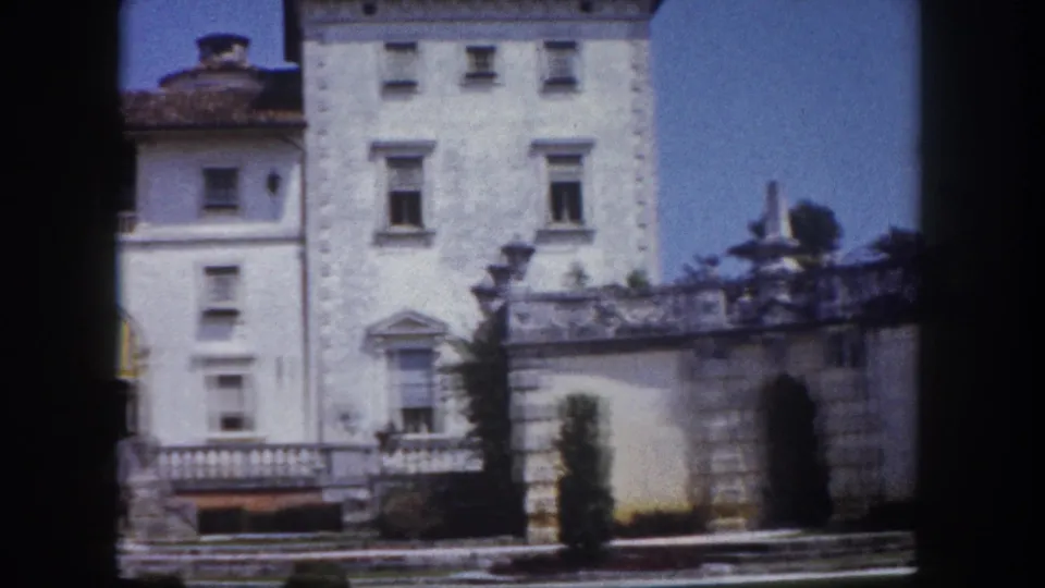 8mm frame circa 1960, Florida: white Mediterranean Revival mansion with central tower or cupola, multiple rows of windows, ornate stone balustrade in foreground, blue sky.