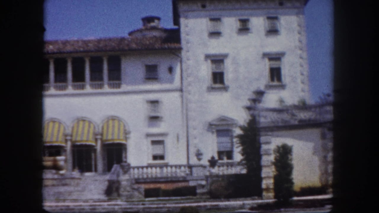 White multi-story Italian Renaissance-style estate building with yellow and green striped arched awnings, stone balustrade terrace and steps, within visible film gate borders.