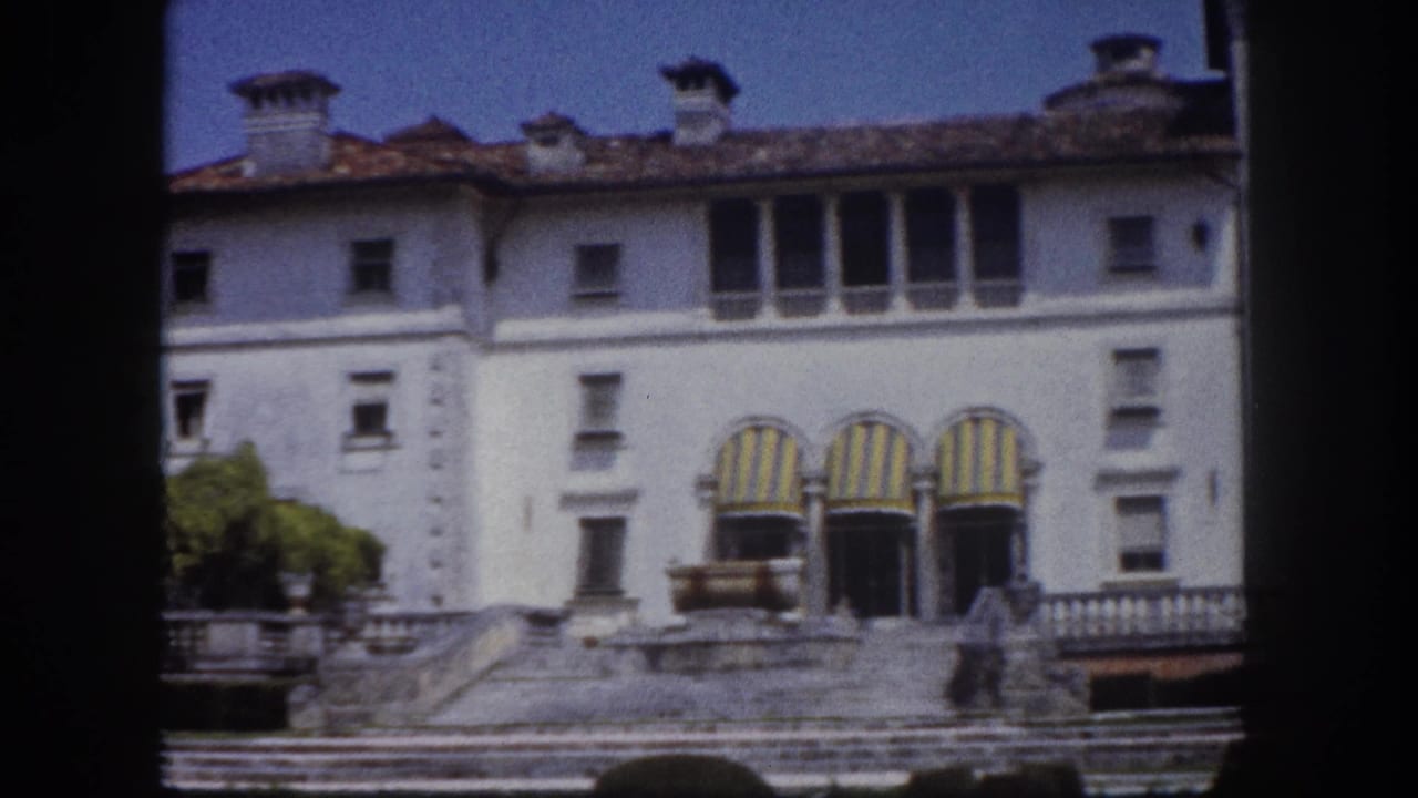 8mm film frame circa 1960 showing a rightward pan across a white Florida estate building, revealing the right wing's striped awnings, tiled roofline, and stone balustrade.