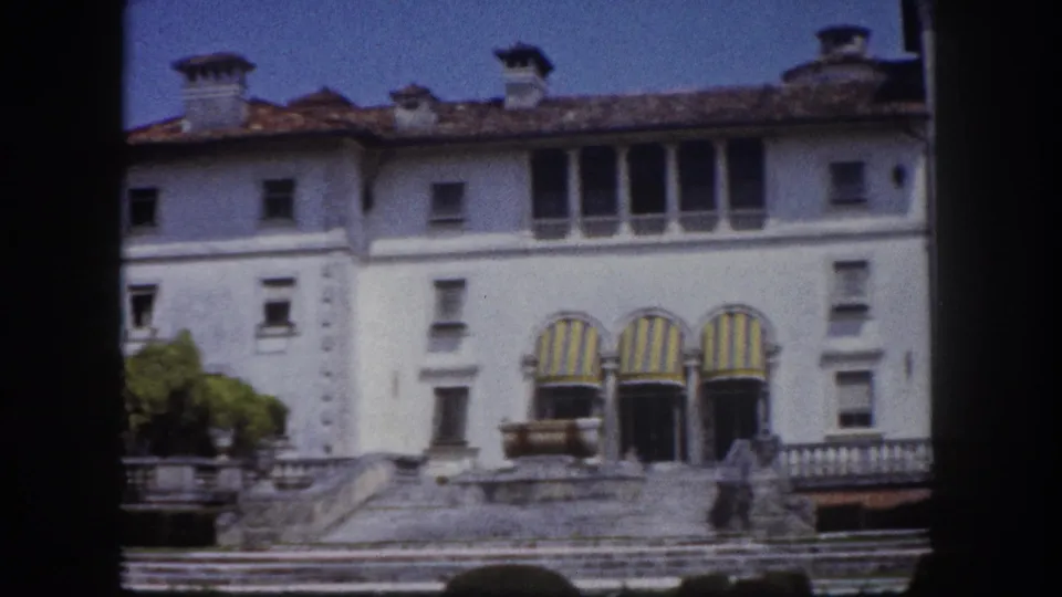 8mm film frame circa 1960 showing a rightward pan across a white Florida estate building, revealing the right wing's striped awnings, tiled roofline, and stone balustrade.