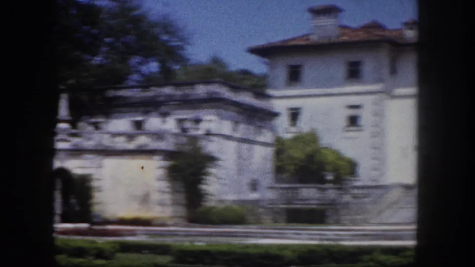 8mm film frame circa 1960 showing the tower and lateral wing of a white Florida estate from garden level, with stone boundary walls and mature shade trees.