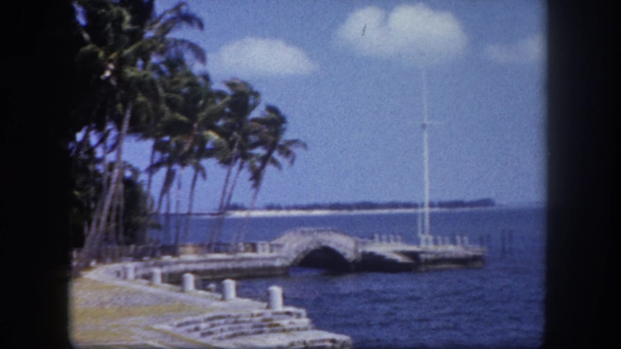 Stone pier with arched bridge and palm trees along a blue bay, 8mm home movie footage.