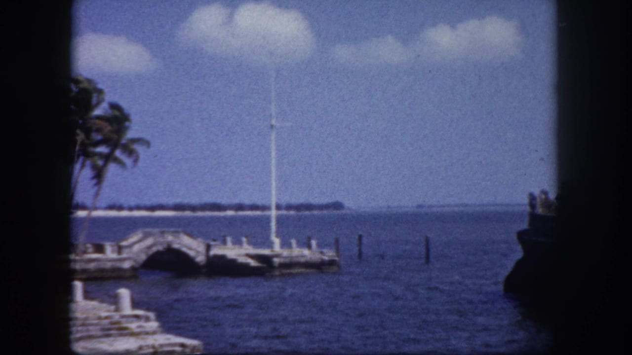 8mm home movie frame circa 1960 showing a stone dock with arched bridge, flagpole, palm trees, and open bay water in the Tampa, Florida area.