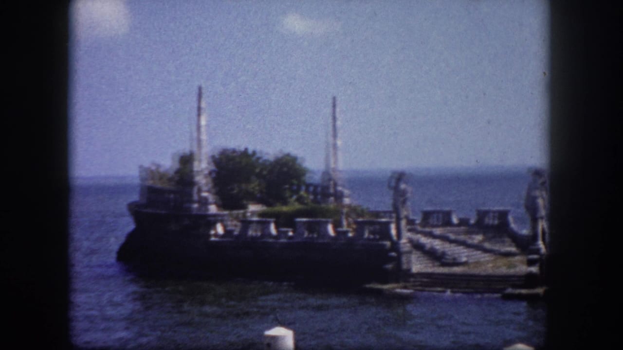 Stone waterfront ruins with tall spires and carved statues extending over calm open water, 8mm home movie frame.