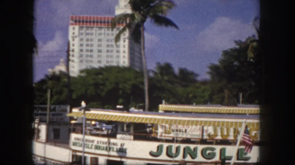 Jungle Cruise excursion boat with large painted green lettering and American flags docked at a Florida waterfront, circa 1960, with a mid-century hotel and palm trees behind.