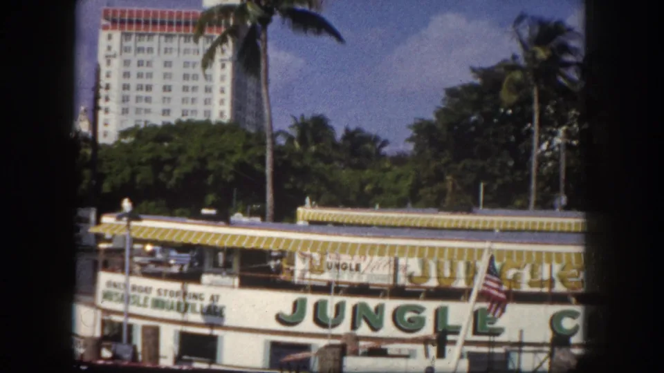 Jungle Cruise excursion boat bow with large painted letters and American flags at a Florida waterfront, circa 1960, hotel visible behind palm trees.