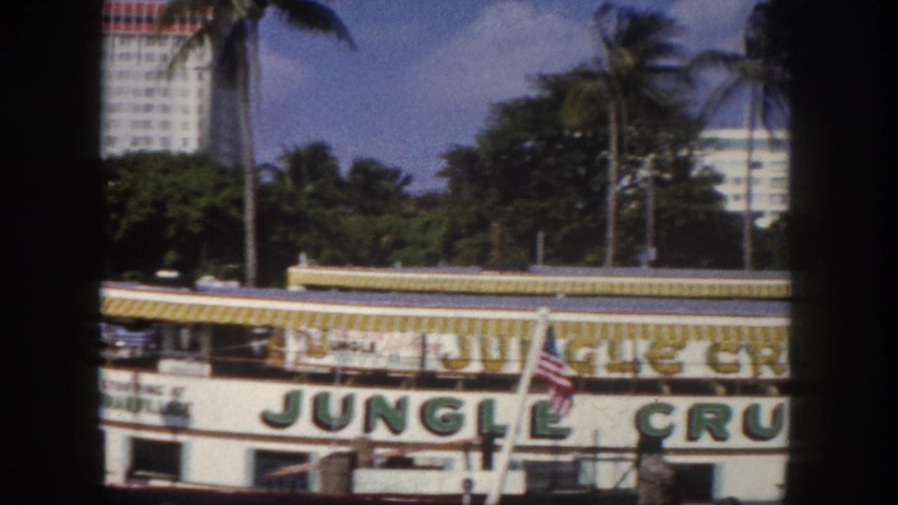 White riverboat with large green Jungle Cruise lettering and yellow-striped awning, palm trees and a white building visible behind.