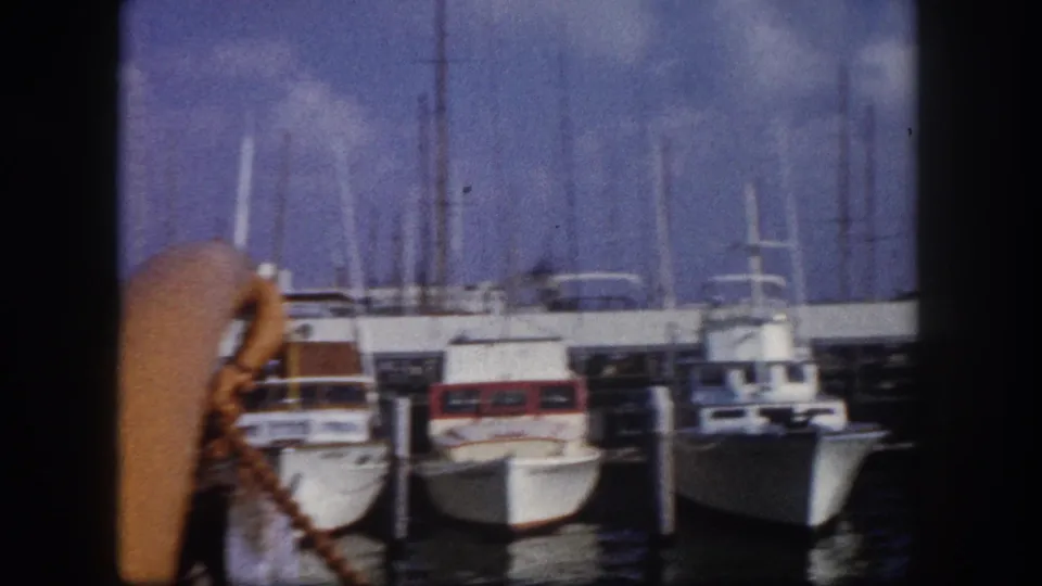 Motorboats moored along dock pilings at a Florida marina circa 1960; an orange rope coil or boat fender is prominent at lower left, with palm trees above.