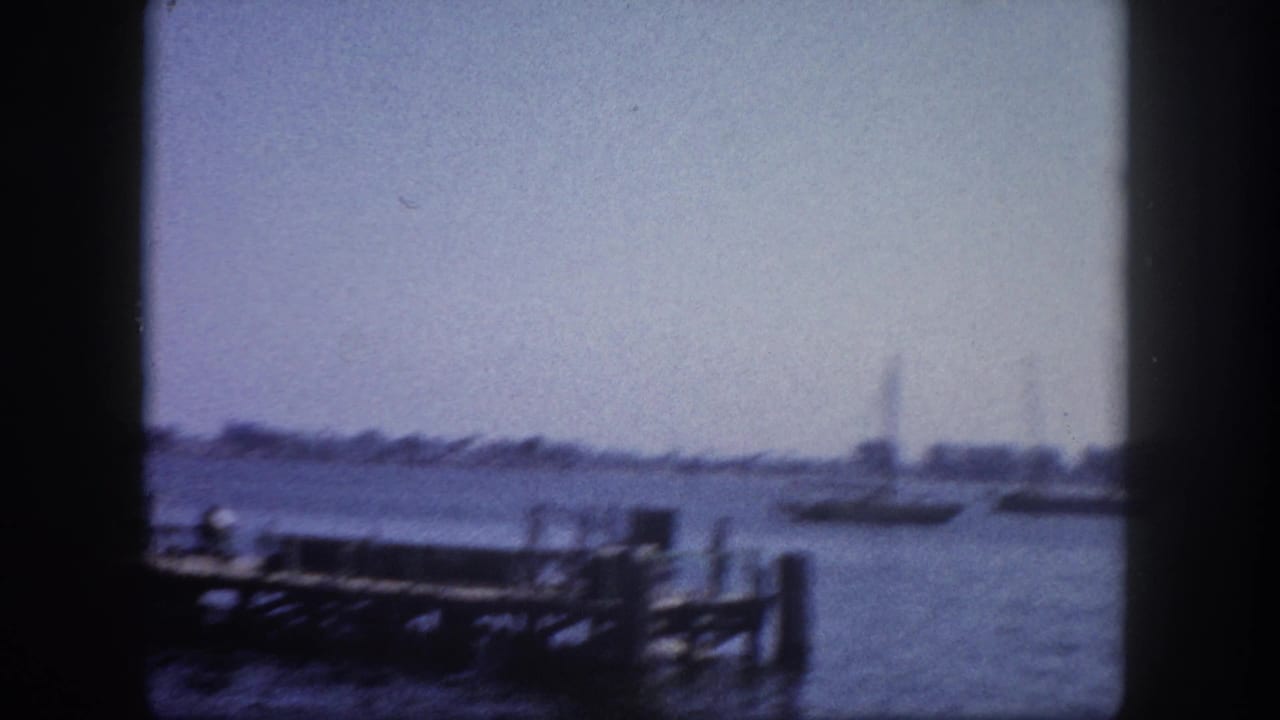 8mm film frame of Tampa Bay waterfront, circa 1960; dock pilings lead into open water with distant vessel silhouettes and treeline visible.