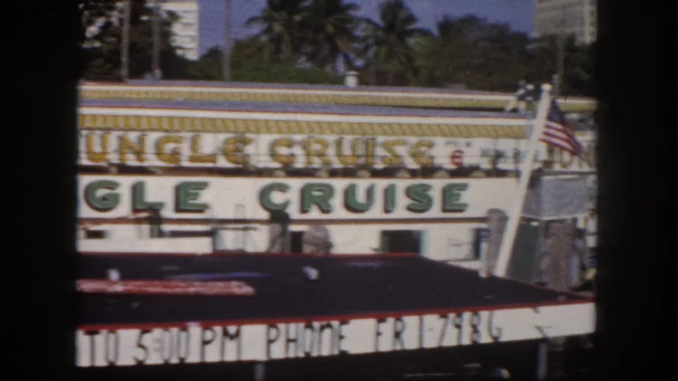 8mm film frame showing a Jungle Cruise waterfront attraction building in Tampa, Florida, circa 1960; American flag, palm trees, and period phone-exchange text visible.