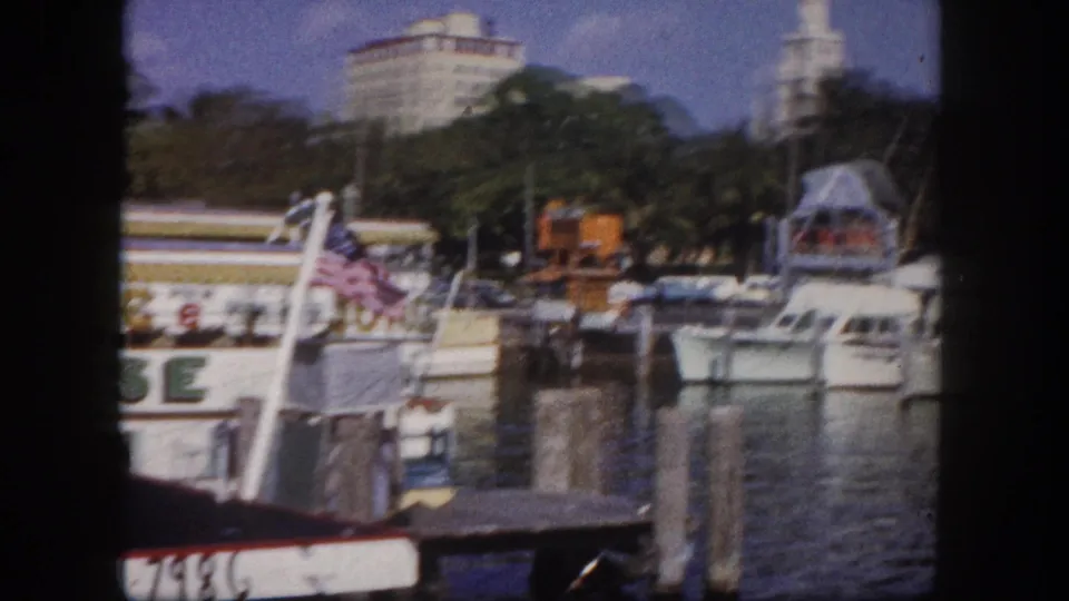 8mm film frame of Tampa marina, circa 1960; white motorboat moored at dock with Jungle Cruise signage, American flag, and a cylindrical tower building in background.