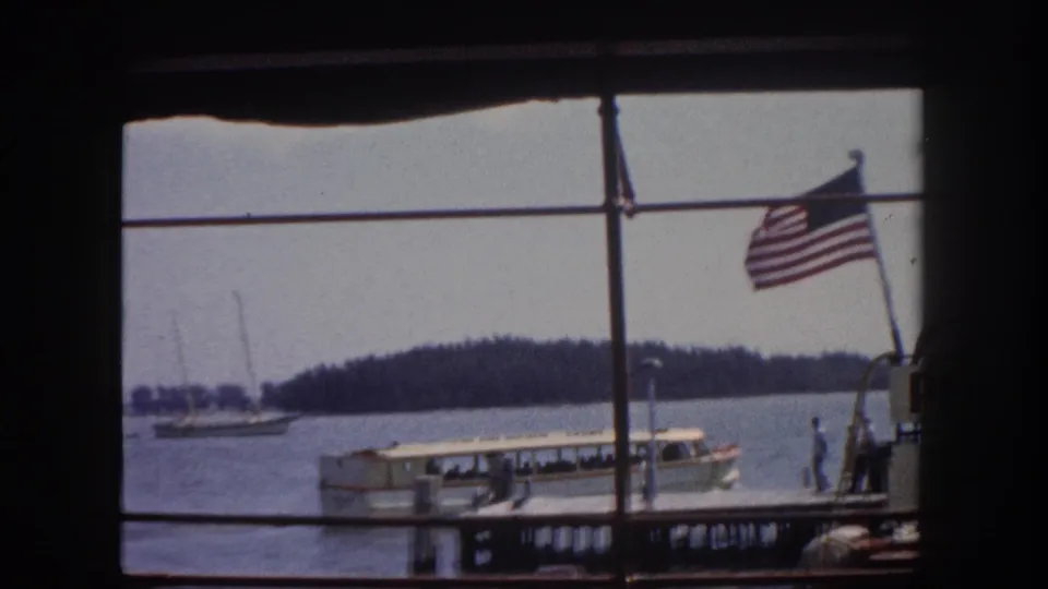 American flag prominently waving, large white excursion boat moored at dock, small figures visible near dock, Tampa Bay marina c.1960, 8mm film.