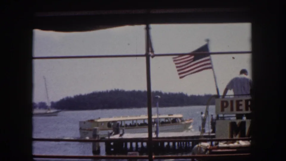 White tour boat at a Florida marina dock, circa 1960, viewed through a window; American flag and partial pier signage visible alongside a silhouetted dock figure.