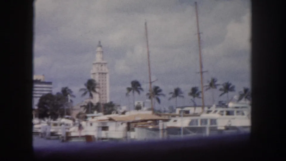 8mm home movie frame c. 1960, Tampa Florida marina with moored yachts, palm trees, and a distant white tower consistent with a civic landmark