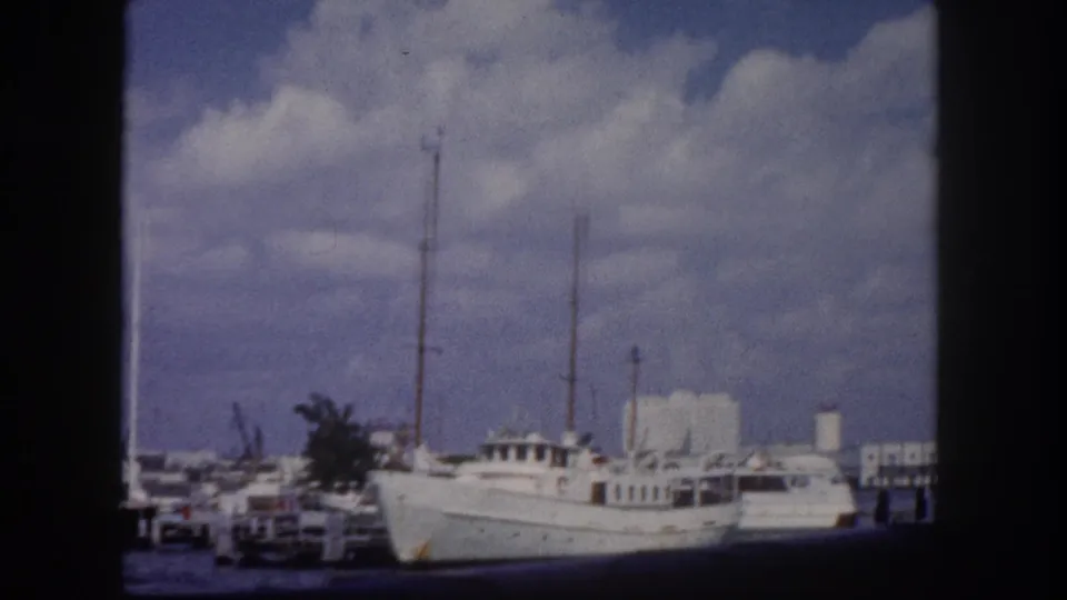 8mm home movie frame c. 1960, large white motor yacht moored at a Florida marina dock with white waterfront buildings and cumulus clouds behind