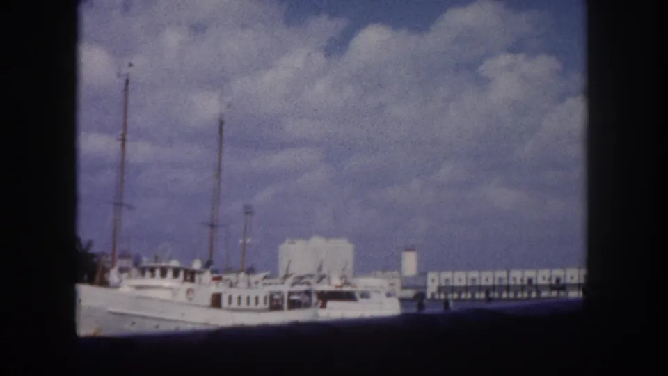 8mm home movie frame c. 1960, same or adjacent large white motor yacht in Florida marina, pier structure and white waterfront buildings behind
