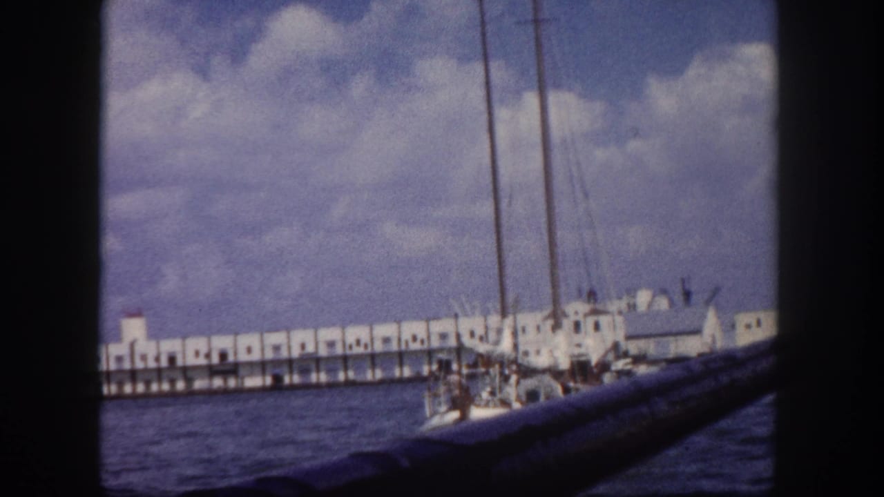 Long white colonnaded pier with repeated arched openings stretching across frame, sailboat masts in foreground, calm harbor water.