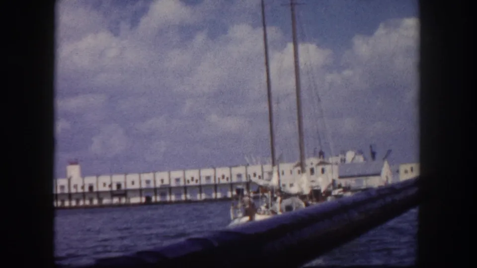 8mm footage circa 1960 with sailboat masts and hull close in frame and the long white pier structure visible behind across the harbor.