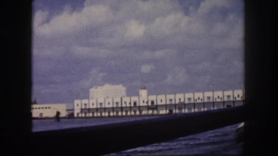 8mm home movie frame, circa 1960, Tampa harbor: long white waterfront shed with repetitive arched bays and a small distant figure, seen from the water.